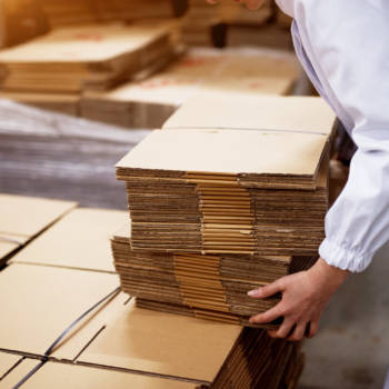 Close up of young female worker picking up stacks of folded cardboard boxes from a bigger stack in factory storage room.